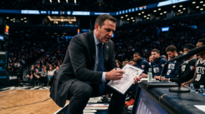 NBA head coach crouched at sideline during a timeout, gesturing strategy to players on the bench under arena spotlights